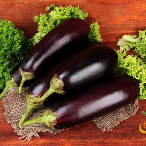 Three glossy dark purple eggplants on burlap surrounded by green lettuce leaves on wooden surface, representing Eggplant - Florida Market seeds.